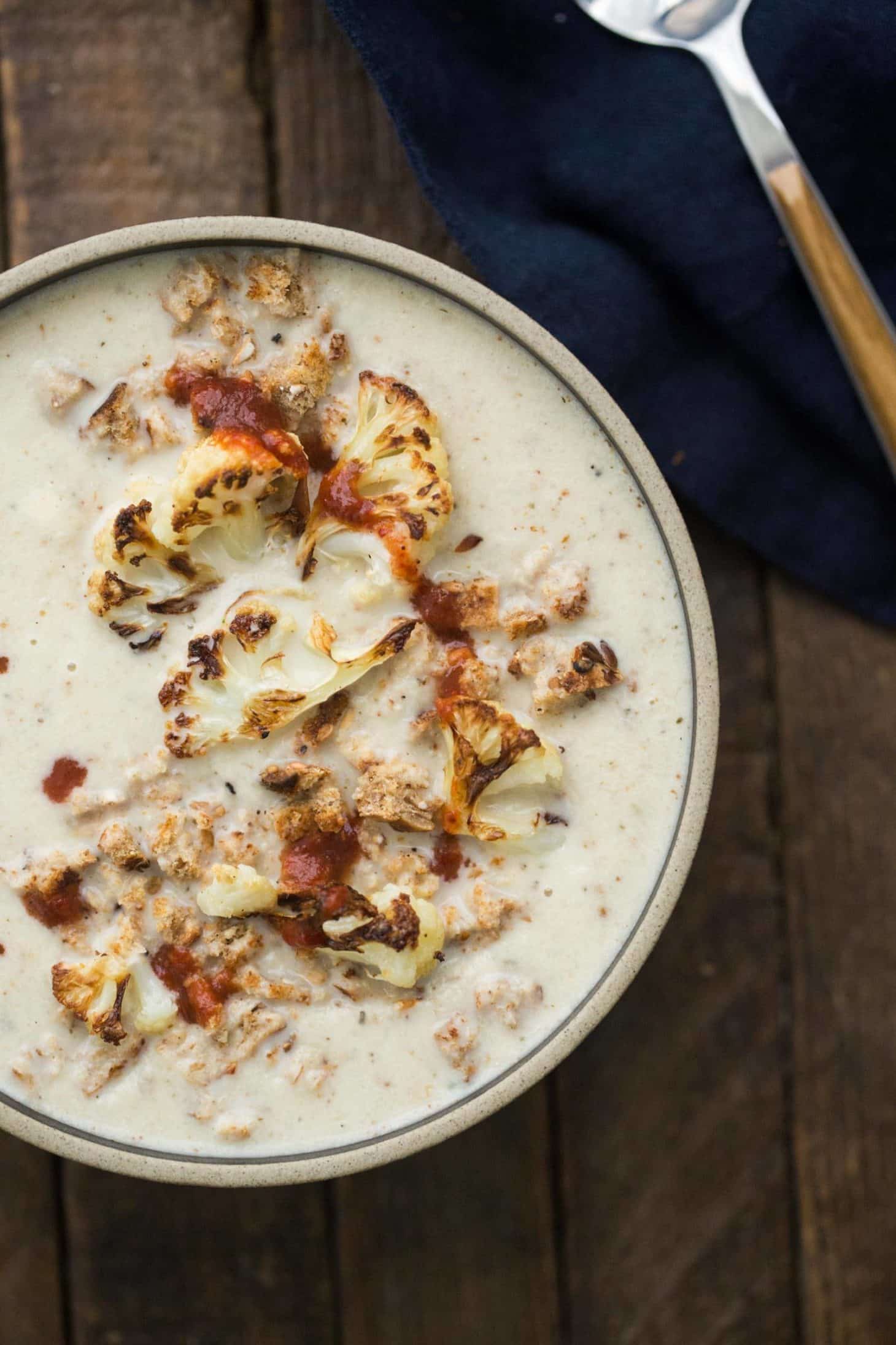 Close-up, overhead photograph of cauliflower soup topped with pieces of roasted cauliflower, breadcrumbs, and hot sauce.