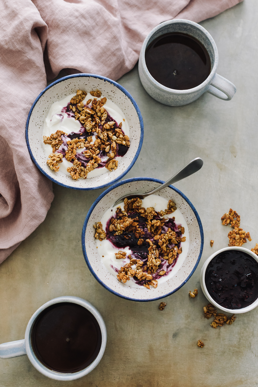 Overhead shot yogurt topped with blueberry jam and granola with cups of coffee and a pink linen.
