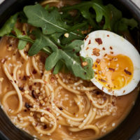 Black bowl filled with noodles, orange broth, arugula, and an egg sliced in half on a light grey background