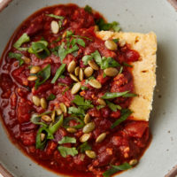 Close-up photo of tempeh chili smothered a piece of cornbread in a grey bowl.