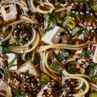 Close-up image of noodles and tofu in a tan broth, scallions, and bok choy