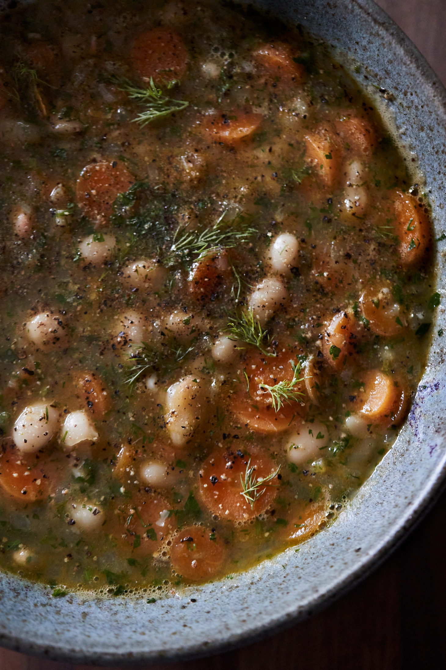 Close-up, overhead photo of a blue ceramic bowl with white beans and carrots in broth with herbs on top.